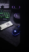 A dark, dimly lit workspace featuring a mechanical keyboard with green backlighting, a smartphone displaying a phone number, a backlit gaming mouse, and a set of headphones. The atmosphere appears to be that of a gaming or tech setup.