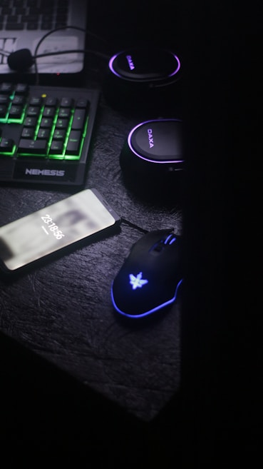 A dark, dimly lit workspace featuring a mechanical keyboard with green backlighting, a smartphone displaying a phone number, a backlit gaming mouse, and a set of headphones. The atmosphere appears to be that of a gaming or tech setup.