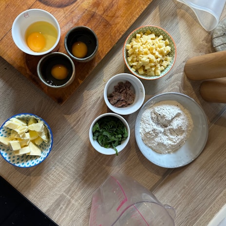 A kitchen workspace featuring an arrangement of baking or cooking ingredients on a wooden surface. There are three containers of eggs, a bowl of diced cheese, a bowl with chocolate or dried fruit, a small bowl of green herbs, a dish of flour with a circular indentation, and a bowl with butter pieces. Wooden rolling pins and a measuring jug can also be seen.