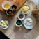 A kitchen workspace featuring an arrangement of baking or cooking ingredients on a wooden surface. There are three containers of eggs, a bowl of diced cheese, a bowl with chocolate or dried fruit, a small bowl of green herbs, a dish of flour with a circular indentation, and a bowl with butter pieces. Wooden rolling pins and a measuring jug can also be seen.