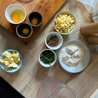 A kitchen workspace featuring an arrangement of baking or cooking ingredients on a wooden surface. There are three containers of eggs, a bowl of diced cheese, a bowl with chocolate or dried fruit, a small bowl of green herbs, a dish of flour with a circular indentation, and a bowl with butter pieces. Wooden rolling pins and a measuring jug can also be seen.