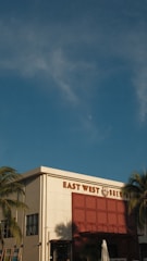 A brewery building with the name 'East West Brewery' displayed prominently. The structure has a modern design with a white facade and large red accent panels. Palm trees frame the sides, and the sky is clear with a few clouds.