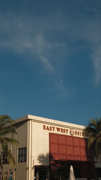 A brewery building with the name 'East West Brewery' displayed prominently. The structure has a modern design with a white facade and large red accent panels. Palm trees frame the sides, and the sky is clear with a few clouds.