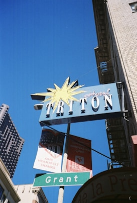A vintage hotel sign with a starburst design at the top, displaying the name 'Hotel Triton' in bold letters on a blue background. Below it, there's a green street sign reading 'Grant' and a couple of banners promoting support for a hospital foundation on a clear blue sky.