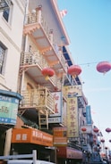 The image features a bustling street scene with buildings adorned with numerous signs in Chinese characters. Red lanterns hang across the street, contributing to a festive and cultural ambiance. The architecture includes balconies and decorative railings, suggesting an East Asian influence.