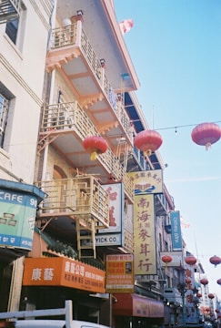 The image features a bustling street scene with buildings adorned with numerous signs in Chinese characters. Red lanterns hang across the street, contributing to a festive and cultural ambiance. The architecture includes balconies and decorative railings, suggesting an East Asian influence.