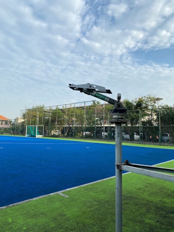 A vibrant blue sports field, possibly for hockey, is bordered by a bright green area. A camera or surveillance device mounted on a metal pole overlooks the scene. In the background, there's a goal net and a perimeter fence with trees and some buildings partially visible under a partly cloudy sky.