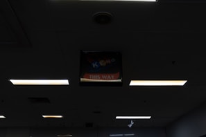 A dimly lit indoor ceiling with several fluorescent light panels. In the center, there is a backlit sign that reads 'KENO THIS WAY' with a partially visible arrow pointing directionally.
