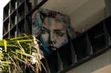 A large, colorful mural of a woman's face is painted on a wall, partially obscured by a balcony structure. The artwork is vibrant, with brush strokes of blue, orange, and red, giving the portrait a dynamic appearance. In the foreground, palm leaves add a natural element against the urban backdrop.