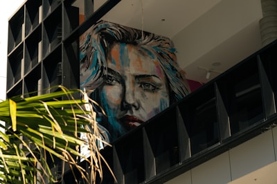 A large, colorful mural of a woman's face is painted on a wall, partially obscured by a balcony structure. The artwork is vibrant, with brush strokes of blue, orange, and red, giving the portrait a dynamic appearance. In the foreground, palm leaves add a natural element against the urban backdrop.