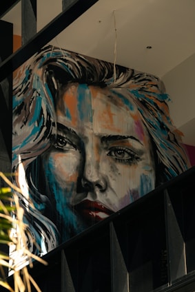 A large, expressive mural portrays a woman's face in a realistic yet artistic style. The painting features dynamic brush strokes with a mix of bright and dark colors, giving it a dramatic appearance. The artwork is situated on an interior wall with visible beams and other architectural elements around it.