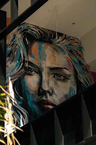 A large, expressive mural portrays a woman's face in a realistic yet artistic style. The painting features dynamic brush strokes with a mix of bright and dark colors, giving it a dramatic appearance. The artwork is situated on an interior wall with visible beams and other architectural elements around it.