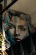 A large, expressive mural portrays a woman's face in a realistic yet artistic style. The painting features dynamic brush strokes with a mix of bright and dark colors, giving it a dramatic appearance. The artwork is situated on an interior wall with visible beams and other architectural elements around it.
