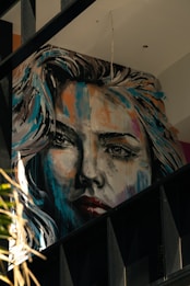 A large, expressive mural portrays a woman's face in a realistic yet artistic style. The painting features dynamic brush strokes with a mix of bright and dark colors, giving it a dramatic appearance. The artwork is situated on an interior wall with visible beams and other architectural elements around it.
