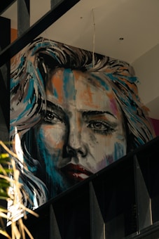 A large, expressive mural portrays a woman's face in a realistic yet artistic style. The painting features dynamic brush strokes with a mix of bright and dark colors, giving it a dramatic appearance. The artwork is situated on an interior wall with visible beams and other architectural elements around it.