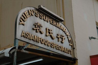 An angled view of a storefront sign featuring large gold letters displaying 'Yuen's Supermarket' and 'Asian Food Specialist' with Chinese characters in between. The sign is mounted on a rectangular structure with a rounded top, beneath which is a metal awning. The background shows a neutral-colored building wall with a mounted light fixture.