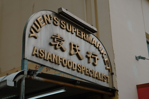 An angled view of a storefront sign featuring large gold letters displaying 'Yuen's Supermarket' and 'Asian Food Specialist' with Chinese characters in between. The sign is mounted on a rectangular structure with a rounded top, beneath which is a metal awning. The background shows a neutral-colored building wall with a mounted light fixture.