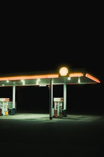 A solitary, well-lit gas station canopy stands alone against a backdrop of complete darkness. The gas pumps beneath the canopy have a soft, ambient glow, while the roof is accentuated by a bold, yellow Shell logo.
