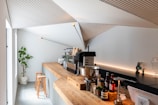 A modern coffee shop interior with a minimalist design. A long wooden counter adorned with various coffee-making accessories extends across the space. A sleek espresso machine is prominently placed on the counter, surrounded by cups and bottles. Natural light enters through a tall window on the left side, highlighting the clean lines and geometric ceiling design. Two wooden stools are neatly positioned by the counter, and a potted plant is placed in the corner, adding a touch of greenery to the scene.
