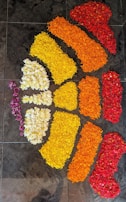 A floral arrangement on a tiled floor, depicting a pattern resembling an abstract design. The arrangement uses flower petals in red, orange, yellow, and white, intricately placed to create vibrant color sections.