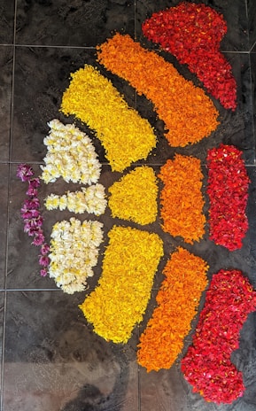 A floral arrangement on a tiled floor, depicting a pattern resembling an abstract design. The arrangement uses flower petals in red, orange, yellow, and white, intricately placed to create vibrant color sections.