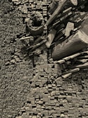 An intricate assemblage of wooden blocks and logs is arranged in a structured pattern. The blocks vary in shape and size, creating a textured, geometric composition. The color palette is monochromatic, emphasizing the texture and forms of the wood.