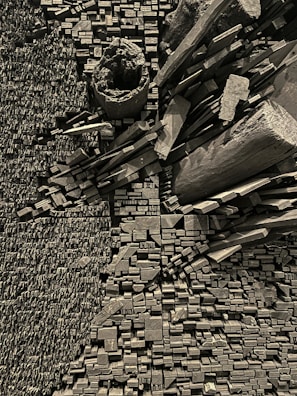 An intricate assemblage of wooden blocks and logs is arranged in a structured pattern. The blocks vary in shape and size, creating a textured, geometric composition. The color palette is monochromatic, emphasizing the texture and forms of the wood.