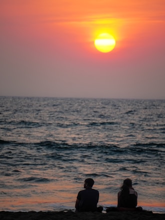 A serene sunset over a vast ocean, with the sun low on the horizon casting warm hues of orange and yellow across the sky. Two people sit quietly on the beach, silhouetted against the tranquil scene, watching the calming waves roll in.