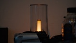 A cylindrical glass object with a warm yellow light emanating from the center. The light source is surrounded by a transparent glass cover, and it's placed next to an electronic device and a plastic bottle on a dark surface.