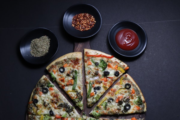 A vegetable pizza is sliced on a wooden board, topped with olives, bell peppers, broccoli, and cheese. Surrounding the pizza are small black bowls containing herbs, chili flakes, and ketchup.