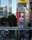 A bustling urban scene with modern buildings, including a large digital billboard displaying an advertisement featuring a woman's face. Busy streets are filled with cars and pedestrians, with an elevated walkway crossing above the traffic. Tall trees line the sidewalk, providing a contrast to the urban structures around.