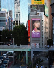A bustling urban scene with modern buildings, including a large digital billboard displaying an advertisement featuring a woman's face. Busy streets are filled with cars and pedestrians, with an elevated walkway crossing above the traffic. Tall trees line the sidewalk, providing a contrast to the urban structures around.