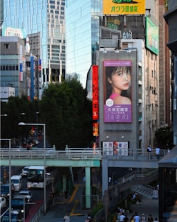 A bustling urban scene with modern buildings, including a large digital billboard displaying an advertisement featuring a woman's face. Busy streets are filled with cars and pedestrians, with an elevated walkway crossing above the traffic. Tall trees line the sidewalk, providing a contrast to the urban structures around.
