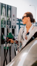 A woman is holding a fuel pump at a gas station. She is wearing sunglasses and a stylish printed outfit. The fuel pump is green and there are multiple fuel dispensers labeled Benzin and Diesel in the background.