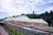 Large greenhouses with beige covers are situated in front of a dense forest under a partly cloudy sky. In the foreground, there are long, horizontal, raised beds or drying racks covered with dark mesh material.