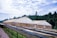 Large greenhouses with beige covers are situated in front of a dense forest under a partly cloudy sky. In the foreground, there are long, horizontal, raised beds or drying racks covered with dark mesh material.