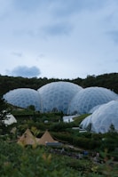Several large geodesic dome structures made of hexagonal panels are set within a lush green landscape. There are trees and shrubbery surrounding and a few tents in the foreground. The sky is mostly overcast with some cloud cover.