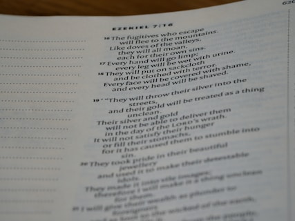A close-up of an open book displaying text from the Bible, specifically Ezekiel 7:16 onwards. The text is printed in a clear, serif font on white paper. The focus is on the written content, with the left side of the page showing dotted lines for notes.