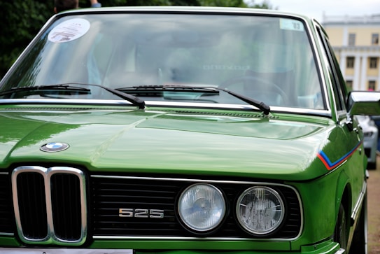 A vibrant green vintage BMW 525 with round headlights and a distinctive grille is prominently featured. The car's polished exterior reflects its surroundings, showcasing its classic design and well-maintained condition. A colorful stripe adorns part of the car, adding a sporty accent.