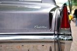 The image features the rear end of a classic Cadillac car, showcasing a dark-colored metallic exterior with chrome details. The tail fin is visible, along with a red tail light. The car's branding is prominently displayed with the Cadillac emblem written in a cursive style on the trunk.