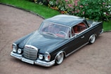 A classic black luxury car with a sleek, polished body and chrome detailing is parked on a gravel path. The car has a vintage design with round headlights and a prominent front grille. The interior is visible through the windows, showing red leather seats. Surrounding the area is a well-maintained garden with colorful flowers and green bushes.