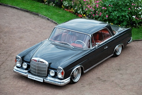 A classic black luxury car with a sleek, polished body and chrome detailing is parked on a gravel path. The car has a vintage design with round headlights and a prominent front grille. The interior is visible through the windows, showing red leather seats. Surrounding the area is a well-maintained garden with colorful flowers and green bushes.
