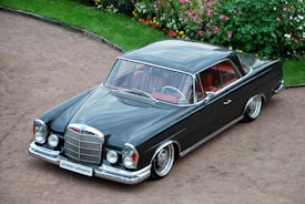 A classic black luxury car with a sleek, polished body and chrome detailing is parked on a gravel path. The car has a vintage design with round headlights and a prominent front grille. The interior is visible through the windows, showing red leather seats. Surrounding the area is a well-maintained garden with colorful flowers and green bushes.