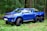 A vibrant blue, custom-built Toyota pickup truck with six wheels is parked on a grass-covered area in a dense forest. The vehicle features oversized off-road tires and chrome detailing, giving it a rugged and powerful appearance.