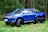 A vibrant blue, custom-built Toyota pickup truck with six wheels is parked on a grass-covered area in a dense forest. The vehicle features oversized off-road tires and chrome detailing, giving it a rugged and powerful appearance.