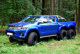 A vibrant blue, custom-built Toyota pickup truck with six wheels is parked on a grass-covered area in a dense forest. The vehicle features oversized off-road tires and chrome detailing, giving it a rugged and powerful appearance.