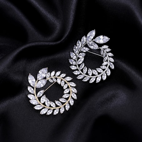 Two elegant, leaf-shaped brooches adorned with sparkling gemstones are positioned on a smooth black, fabric background. The brooches are arranged in a circular pattern, with each leaf-like piece crafted to resemble a delicate branch of foliage.