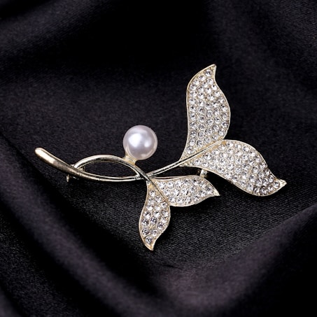 A decorative brooch featuring a stylized leaf design embellished with small, shimmering crystals. The centerpiece is a single lustrous pearl, accentuating the elegant and luxurious appearance. The brooch rests on a backdrop of smooth, black fabric, creating a contrast that highlights its intricate details.