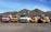 Five trucks are parked in a row on a dusty ground against a backdrop of mountainous terrain. The trucks are of different colors and types, including cement mixers and loaders, with industrial buildings in the background.