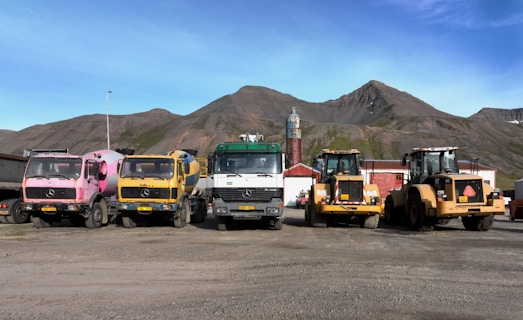 Five trucks are parked in a row on a dusty ground against a backdrop of mountainous terrain. The trucks are of different colors and types, including cement mixers and loaders, with industrial buildings in the background.